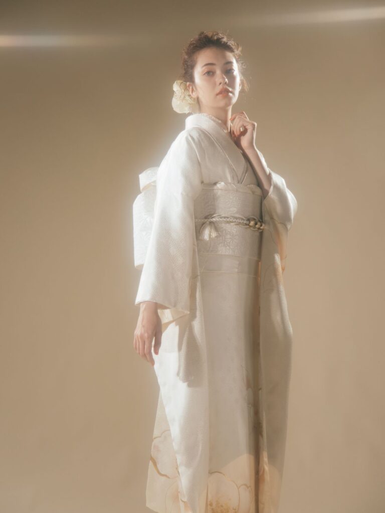 ソフトフォーカスで撮影された、淡い色合いの上品な着物を着た女性。繊細な模様があしらわれた着物に、白い花をあしらったアップヘアを合わせている。背景は温かみのある落ち着いた色調で、片手を顎に当て、思慮深いポーズをとっている。.