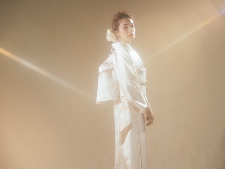 柔らかな金色の照明の下、白い着物に帯を締めた女性が立っている。髪は花飾りで丁寧にまとめられ、温かみのあるベージュの背景が、穏やかで優雅な雰囲気を醸し出している。彼女は肩越しに少し視線を向けている。.