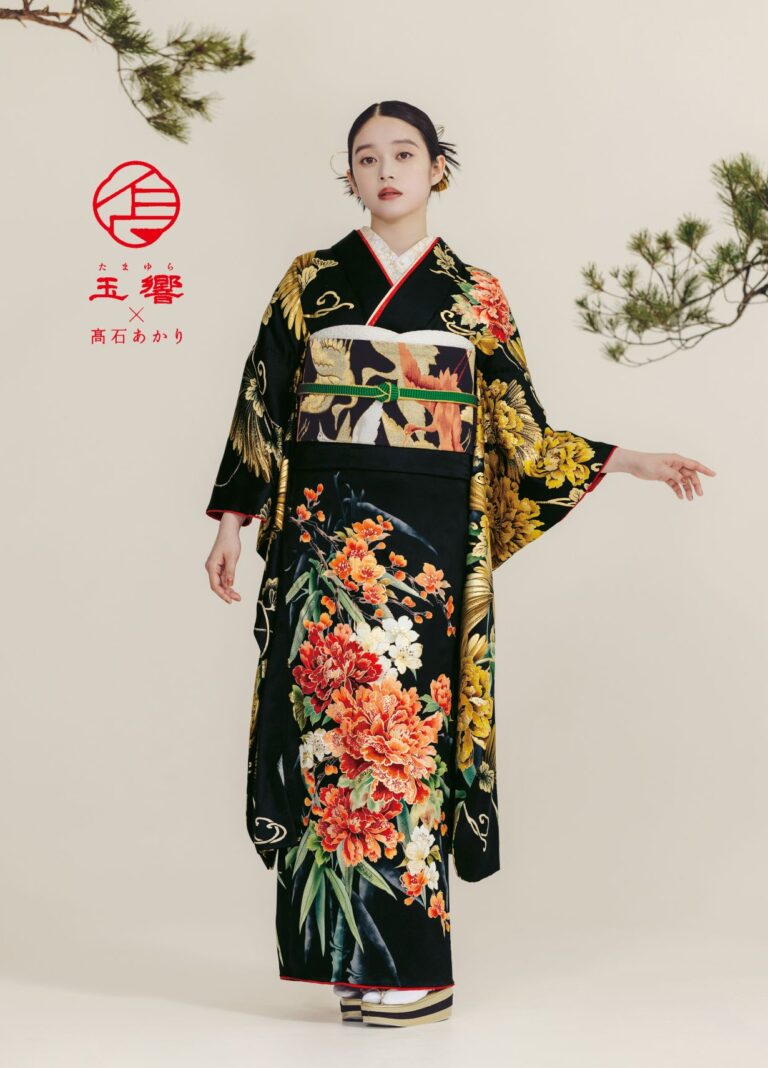 ベージュを背景に、鮮やかな花と鶴の模様が描かれた黒い着物を着た女性が立っている。髪を後ろで結び、穏やかな表情で前を向いている。松の枝が上方を縁取っている。左側に赤い日本語の文字。.