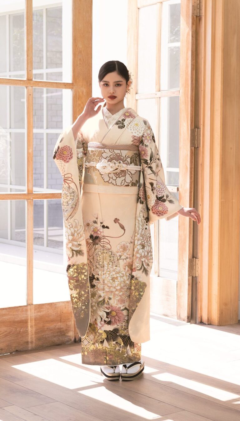 木枠の大きなガラス戸のそばに、花柄の着物を着た女性が優雅に立っている。太陽の光が差し込み、明るいフローリングの床に影を落としている。彼女の背後には木製のキャビネットと階段が見える。.