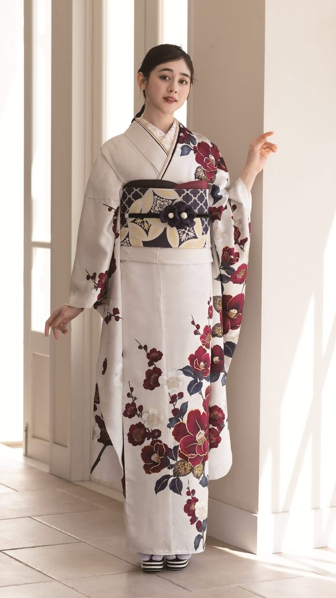 室内で背の高い窓際に立つ女性。赤と黒の花柄が入った白い着物を着て、幅広の飾り帯を締めている。落ち着いた表情でカメラを見つめ、黒髪をきれいに整え、横から陽光が差し込む。.