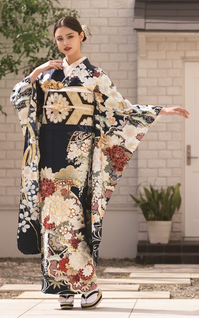 モダンな白いレンガ造りの家の外に、濃い花柄の袖の広い着物を着て金の帯を締めた女性が立っている。両手を広げて優雅にポーズをとっている。明るく日当たりの良い場所で、近くには小さな木と植物がある。.