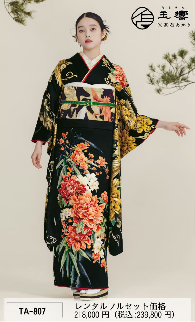 赤、金、オレンジ色の鮮やかな花柄が施された黒い着物を着た女性が立っている。その着物は精巧な模様が特徴で、金と黒を基調とした幅広の帯と、長くふわりと広がる袖がついている。彼女は、淡い色の無地の背景を背にポーズをとっている。.